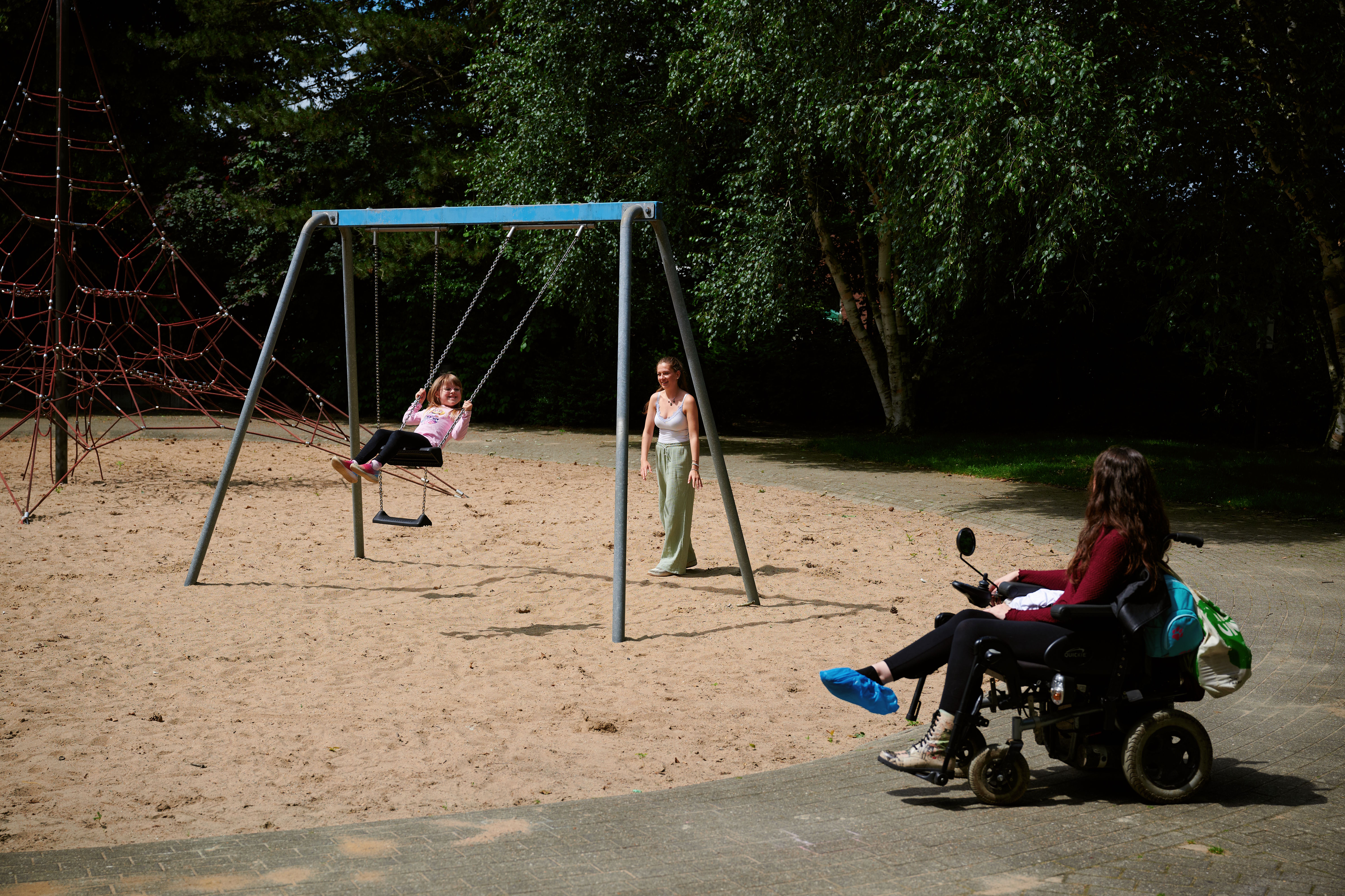 Auf einem Spielplatz wird ein Kind wird von einen jungen Frau angeschaukelt. Daneben sitzt im Rollstuhl die Mutter des Kindes.